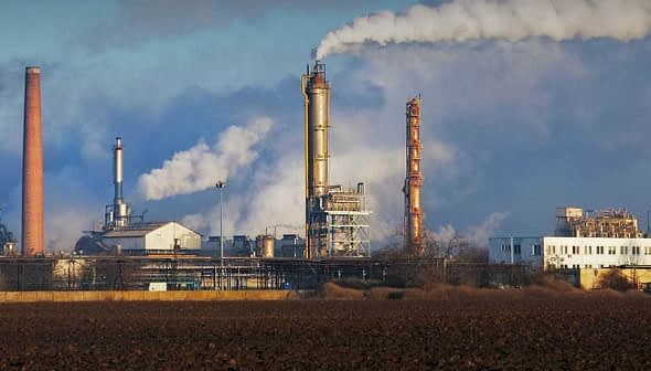 Industrial complex featuring multiple smoke stacks emitting white smoke against a blue sky. - Olive Oil Times