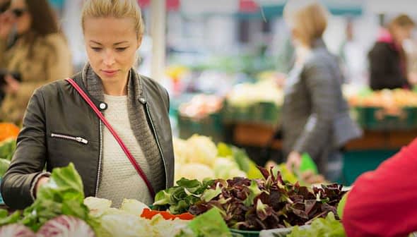 Woman examining fresh vegetables at a market stall surrounded by other shoppers. - Olive Oil Times