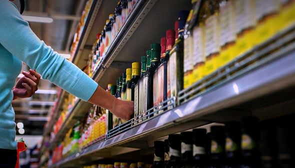 Individual reaching for a bottle of olive oil on a grocery store shelf with various products. - Olive Oil Times