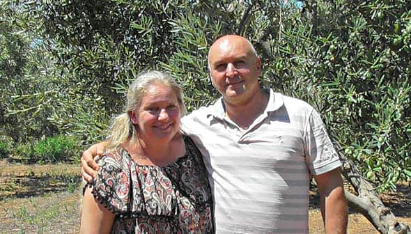 A couple standing together in an olive grove, smiling at the camera. - Olive Oil Times