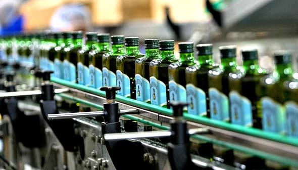 Row of green glass bottles filled with olive oil on a production line in a factory. - Olive Oil Times