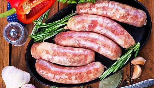 Five raw sausages arranged on a black platter with herbs and garlic nearby. - Olive Oil Times