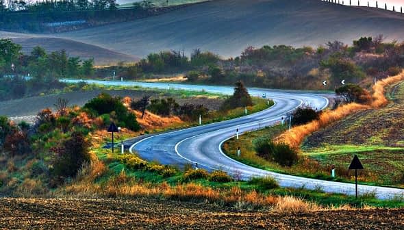 A winding road through a rural landscape with hills and vegetation on either side. - Olive Oil Times