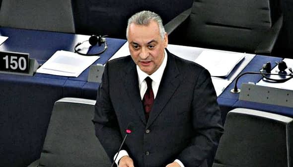 A man speaking at a podium in a legislative assembly with numbered seats visible in the background. - Olive Oil Times