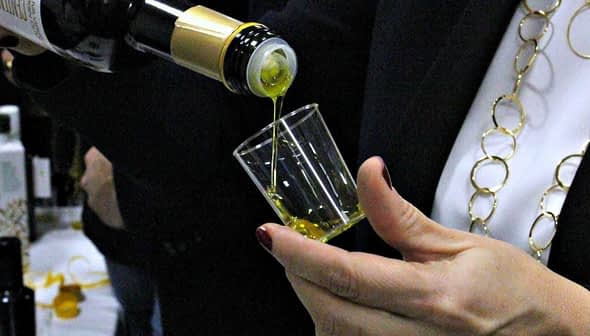 A person pouring olive oil from a bottle into a small glass cup during a tasting event. - Olive Oil Times