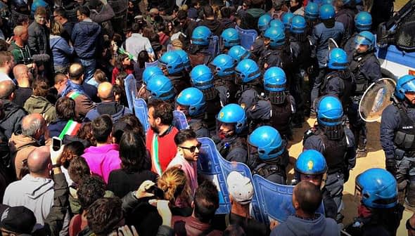 Crowd of people gathered at a demonstration with police officers in riot gear and a police vehicle nearby. - Olive Oil Times