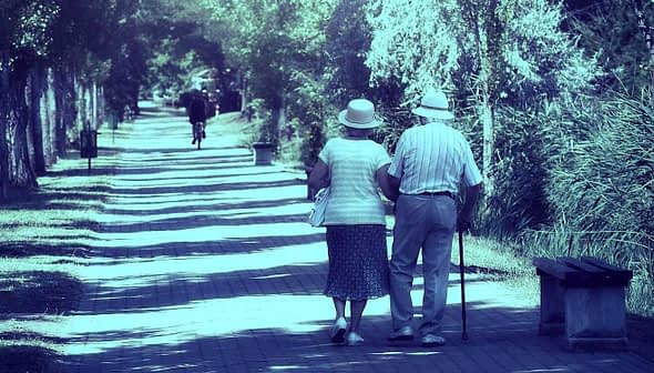 An elderly couple walking together on a tree-lined path, both wearing hats and holding hands. - Olive Oil Times