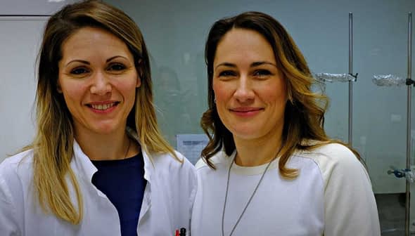 Two women smiling in a laboratory environment, one wearing a lab coat and the other in a white sweater. - Olive Oil Times