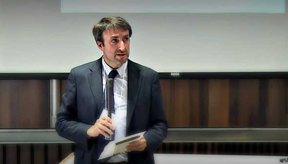 A man in a suit holding a notepad while speaking in front of an audience. - Olive Oil Times