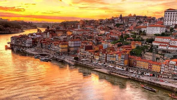 A panoramic view of Porto's riverfront at sunset with colorful buildings along the water's edge. - Olive Oil Times