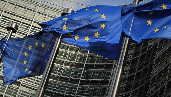 Multiple European Union flags waving in front of a modern glass building. - Olive Oil Times