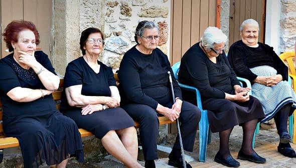 Six elderly women dressed in black and gray sitting on a bench in a casual setting. - Olive Oil Times