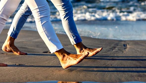 Two individuals walking barefoot along the shoreline with waves in the background. - Olive Oil Times