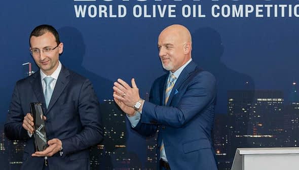 Two men at the World Olive Oil Competition award ceremony, one holding a trophy and the other applauding. - Olive Oil Times