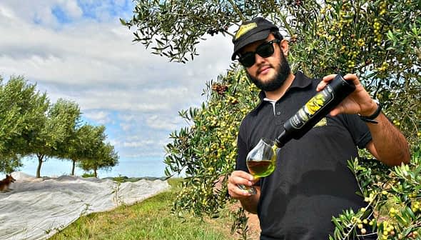 Man in sunglasses pouring olive oil from a bottle into a glass while standing among olive trees. - Olive Oil Times
