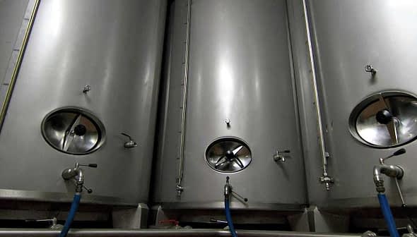 Three stainless steel fermentation tanks with circular openings and valves. - Olive Oil Times