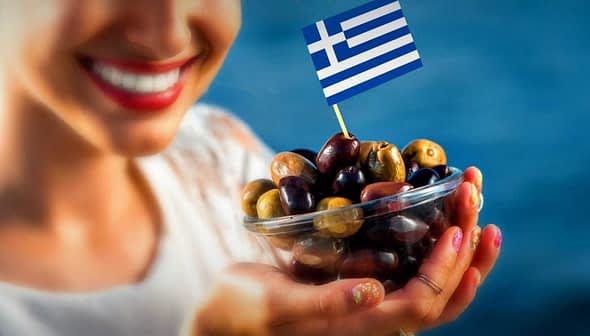 A woman smiling while holding a bowl of assorted olives topped with a small Greek flag. - Olive Oil Times