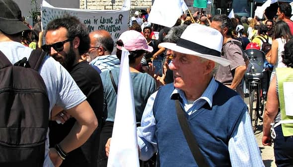An older man wearing a white hat and holding a small flag at a crowded public event. - Olive Oil Times