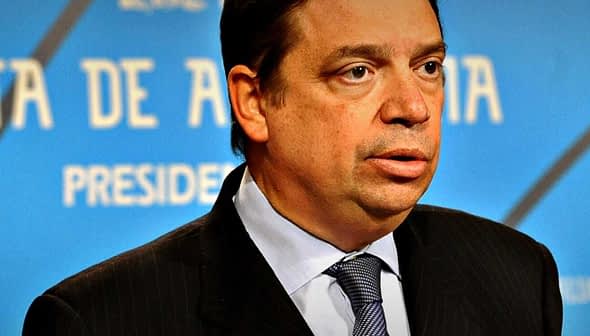 Man in a dark suit and tie speaking at a press conference with a blue background. - Olive Oil Times