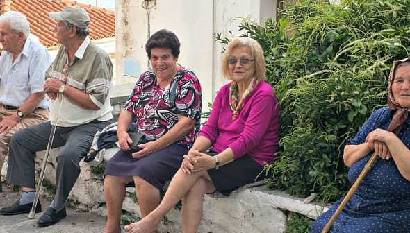 Five elderly individuals sitting on a stone wall in a village setting, with greenery in the background. - Olive Oil Times
