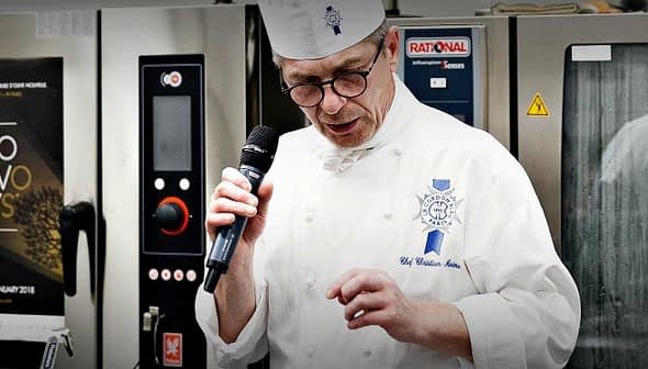 Chef in a white uniform and hat speaking into a microphone at a culinary event. - Olive Oil Times