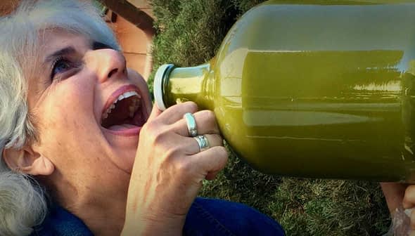 Woman with gray hair drinking from a large green glass bottle outdoors. - Olive Oil Times
