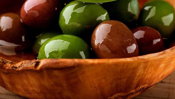 A wooden bowl filled with a variety of green and brown olives resting in oil. - Olive Oil Times