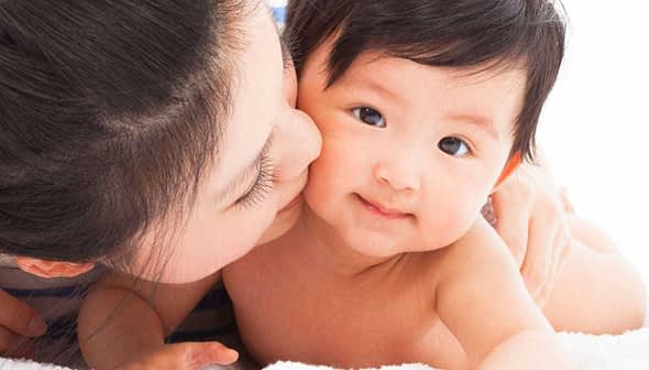 A mother kissing her baby on a soft surface, both looking at the camera. - Olive Oil Times