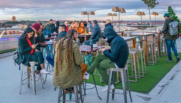 Group of people sitting and standing at a rooftop bar with tables and chairs. - Olive Oil Times