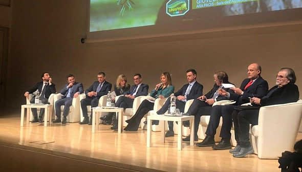 A panel of speakers seated on stage during a conference event, discussing various topics. - Olive Oil Times