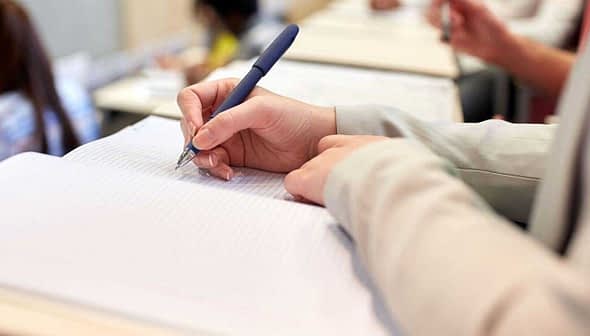 A person holding a pen and writing in a notebook during a classroom setting. - Olive Oil Times