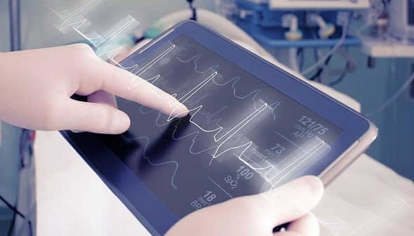 Healthcare professional interacting with a tablet displaying heart rate and vital signs data. - Olive Oil Times