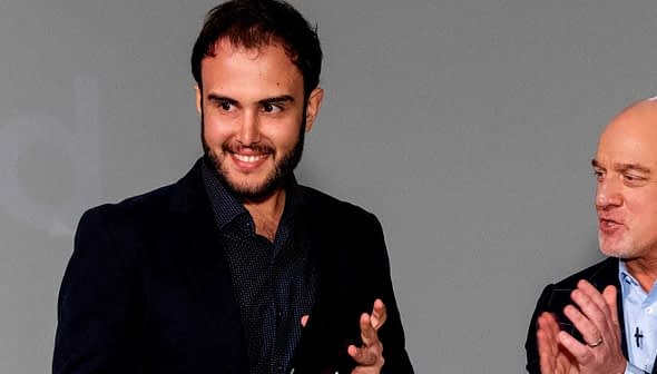 Two men standing on stage, one smiling and clapping while the other speaks. - Olive Oil Times