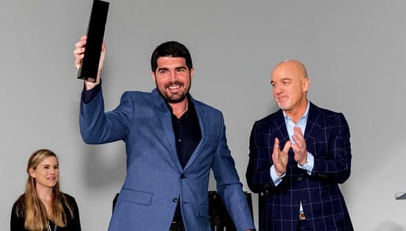 A man in a blue suit holding an award while two others applaud in the background. - Olive Oil Times