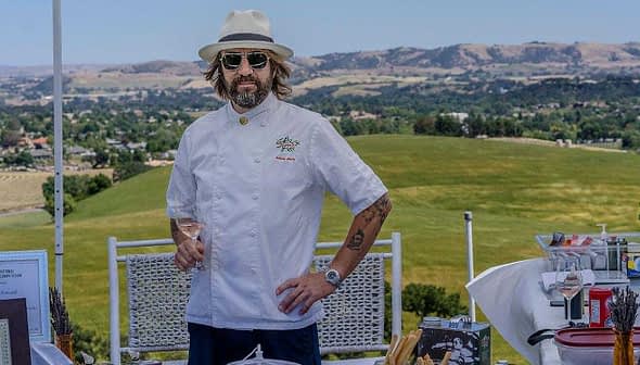 Chef wearing a white coat, hat, and sunglasses standing outdoors with a scenic background. - Olive Oil Times