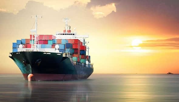 Cargo ship loaded with containers sailing on water during sunset with colorful sky. - Olive Oil Times