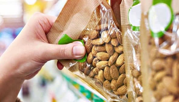 A hand reaching for a transparent package of almonds on a store shelf. - Olive Oil Times