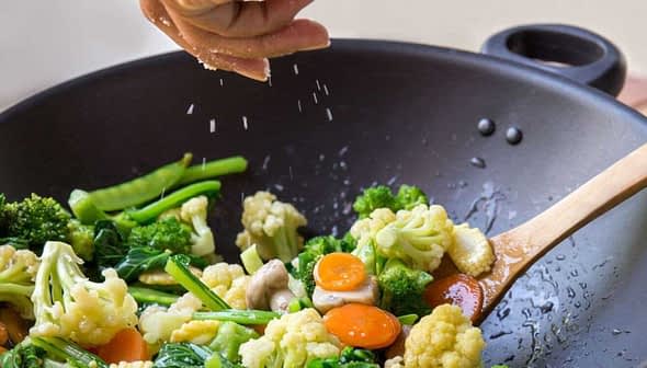 Person sprinkling salt over a mix of vegetables in a black wok with a wooden spoon. - Olive Oil Times