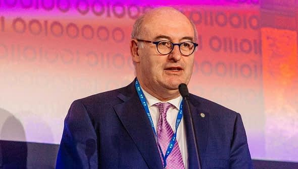 Phil Hogan speaking at a podium during an event with a microphone in front of a colorful background. - Olive Oil Times