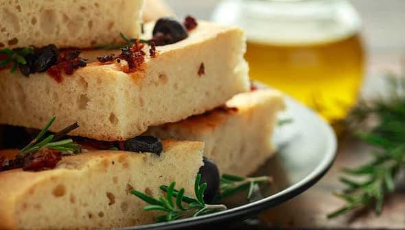 Slices of focaccia bread topped with olives and herbs on a black plate. - Olive Oil Times