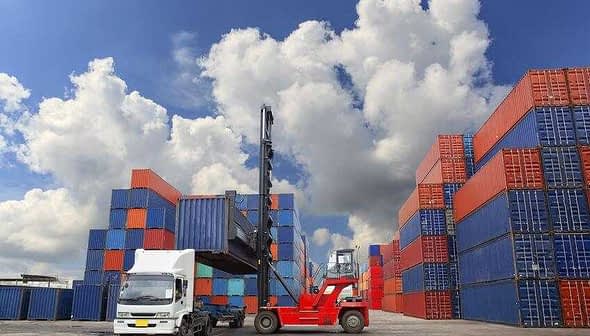 Uma empilhadeira levantando um contêiner de transporte em uma área de armazenamento cercada por contêineres empilhados. Olive Oil Times