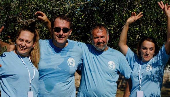 Four individuals wearing blue t-shirts posing together outdoors under olive trees. - Olive Oil Times