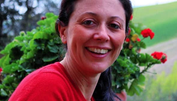 A woman smiling while holding a pot of flowers with red blooms outdoors. - Olive Oil Times