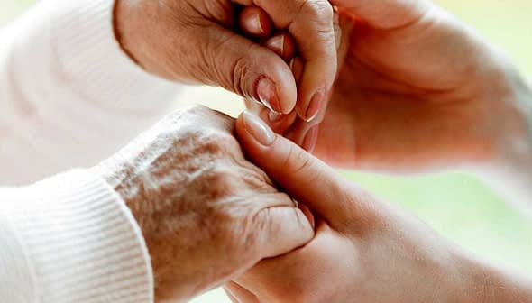 An elderly person's hand being held by a caregiver's hand in a supportive gesture. - Olive Oil Times