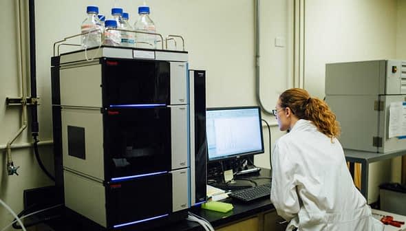 Laboratory technician in a white coat operating analytical equipment and reviewing data on a computer screen. - Olive Oil Times
