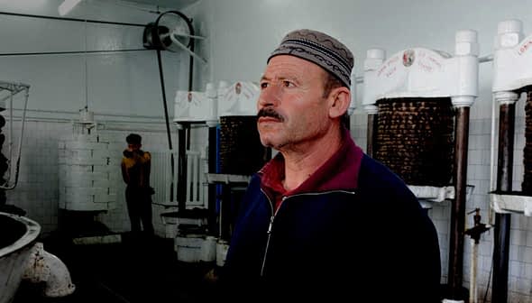 Man wearing a traditional hat standing inside an oil pressing facility with machinery in the background. - Olive Oil Times