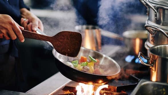 Chef using a wooden spatula to stir colorful vegetables in a frying pan over a flame. - Olive Oil Times