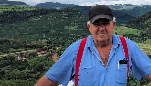 Man wearing a blue shirt and red suspenders standing outdoors with green hills in the background. - Olive Oil Times
