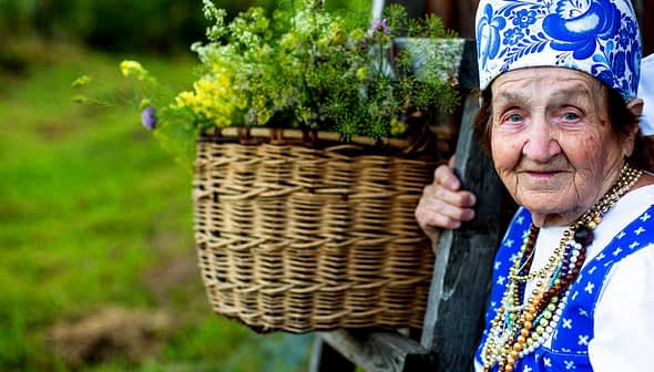 Elderly woman wearing traditional clothing and a headscarf, holding a basket of flowers. - Olive Oil Times