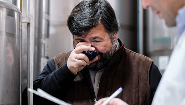 Man smelling olive oil from a small cup while another person observes in the background. - Olive Oil Times